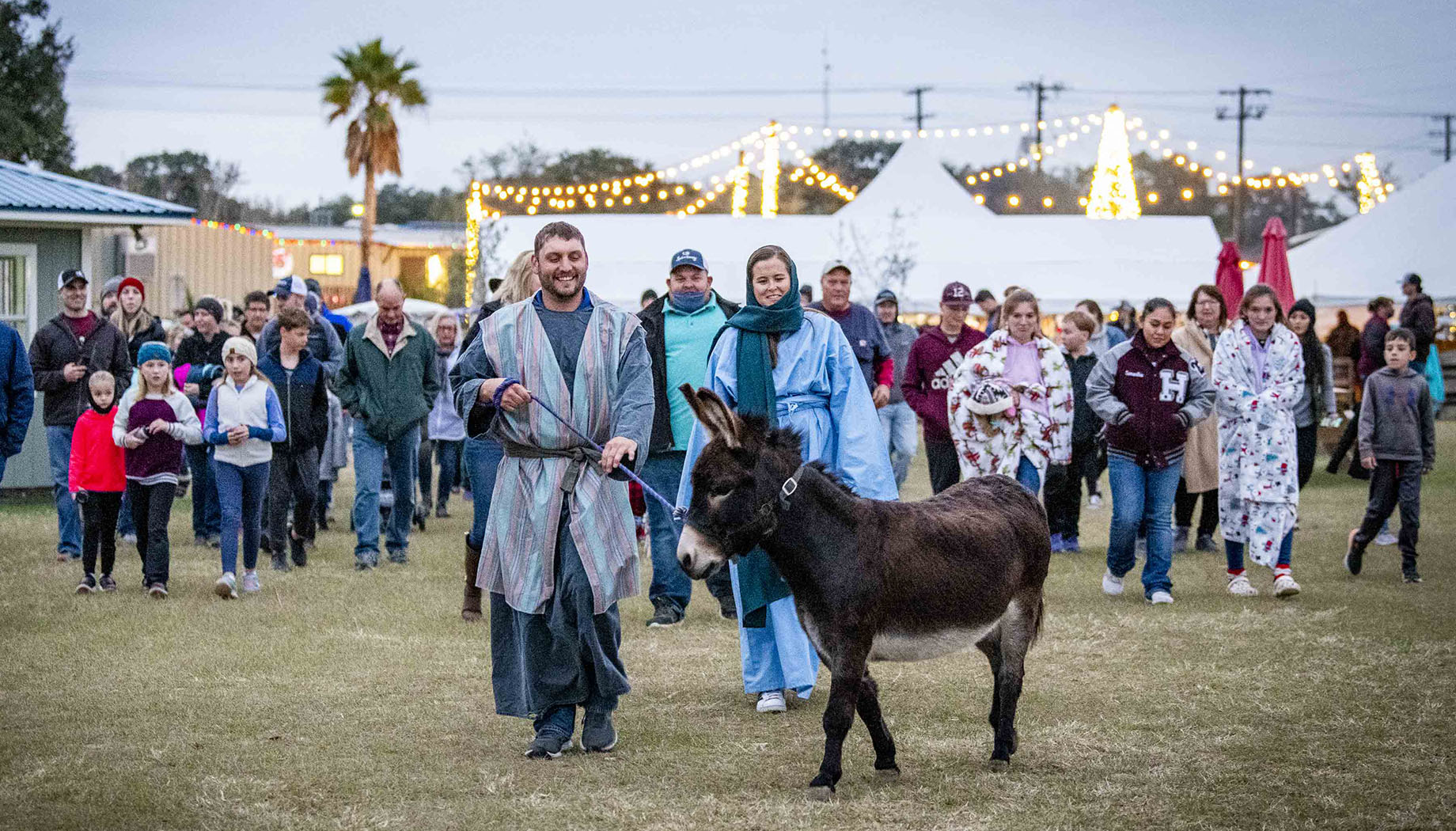 BLOG A group of people, including a man and woman leading a donkey, walk together outdoors in a festive setting with lights in the background.
